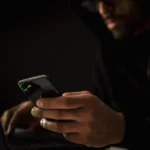Person using a smartphone and laptop in a dark room representing AI-generated phishing attacks
