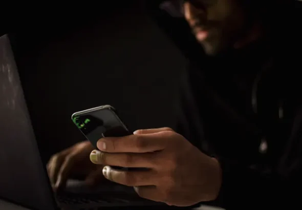 Person using a smartphone and laptop in a dark room representing AI-generated phishing attacks
