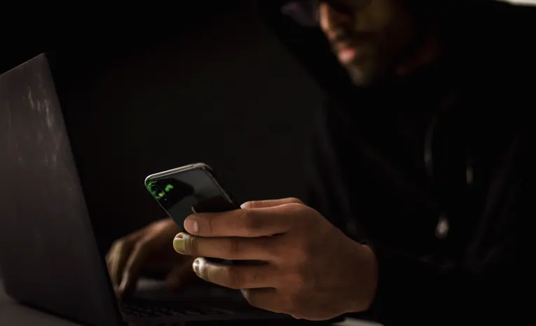 Person using a smartphone and laptop in a dark room representing AI-generated phishing attacks